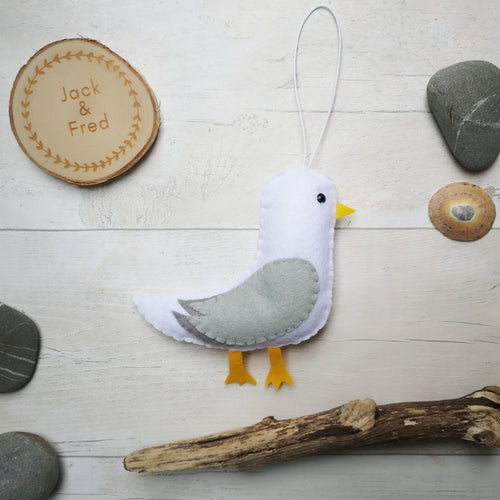handmade felt seagull decoration made from white, grey and yellow felt pictured with pebbles, a shell and driftwood for a beach theme.